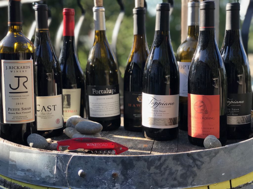 Bottles of Petite Sirah in the sun on a wine barrel with a red corkscrew 