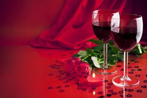 Two glasses of red wine on a red background with red roses and a heart shaped candle