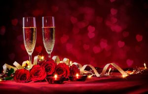 Two flutes filled with sparkling wine and set among red roses, gold ribbon and votive candles.