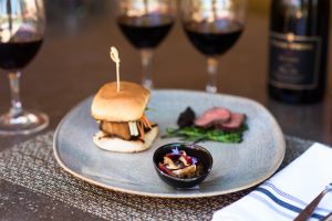 Image of a plate with a slider and some other food with three glass of red wine in the background.