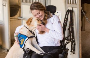 Young woman in a wheelchair cuddling with a Canine Companion yellow lab with a working cap on.