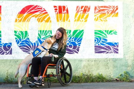 A young woman in a wheelchair with a Canine Companion yellow lab with their paws on her lap and faces next to each other. Large LOVE poster behind them.