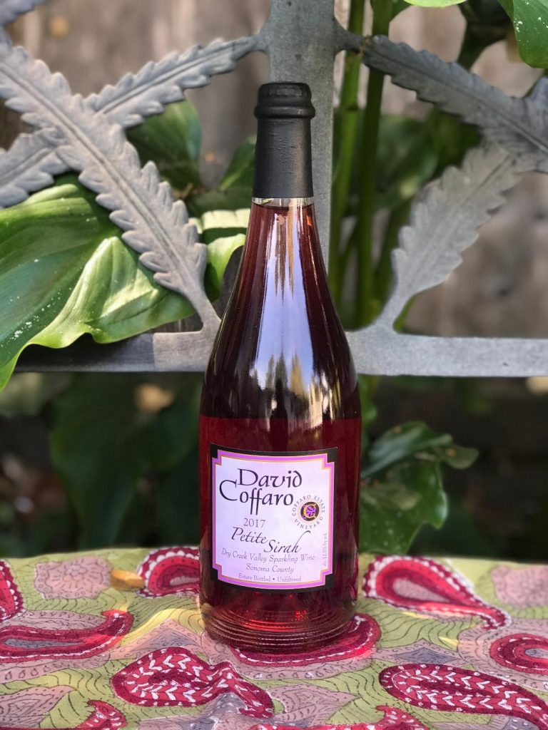 Bottle of David Coffaro Sparkling Peite Sirah on a bench with plant in background