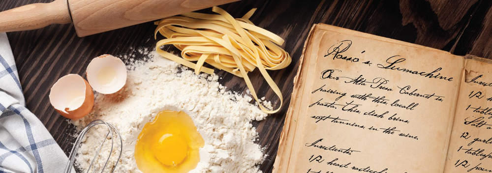 Handwritten recipe in an old book, and to the left side is a pile of flour with a raw egg in a well in the flour, two halves of an empty egg shell, a whisk, some fresh pasta, a checked towel and a rolling pin.