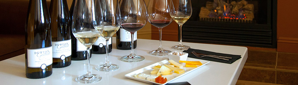 A platter of assorted cheeses, five wine glasses with a small amount of wine in each glass, and wine bottles behind each wine glass.