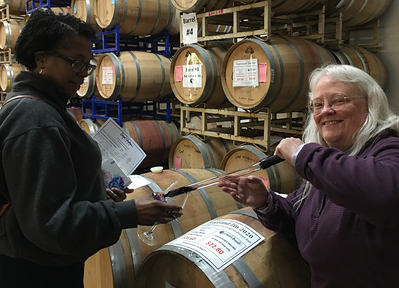 Woman holding wine glass in front of a wine barrel. Carol Shelton is using a wine thief to fill the woman's glass with red wine.