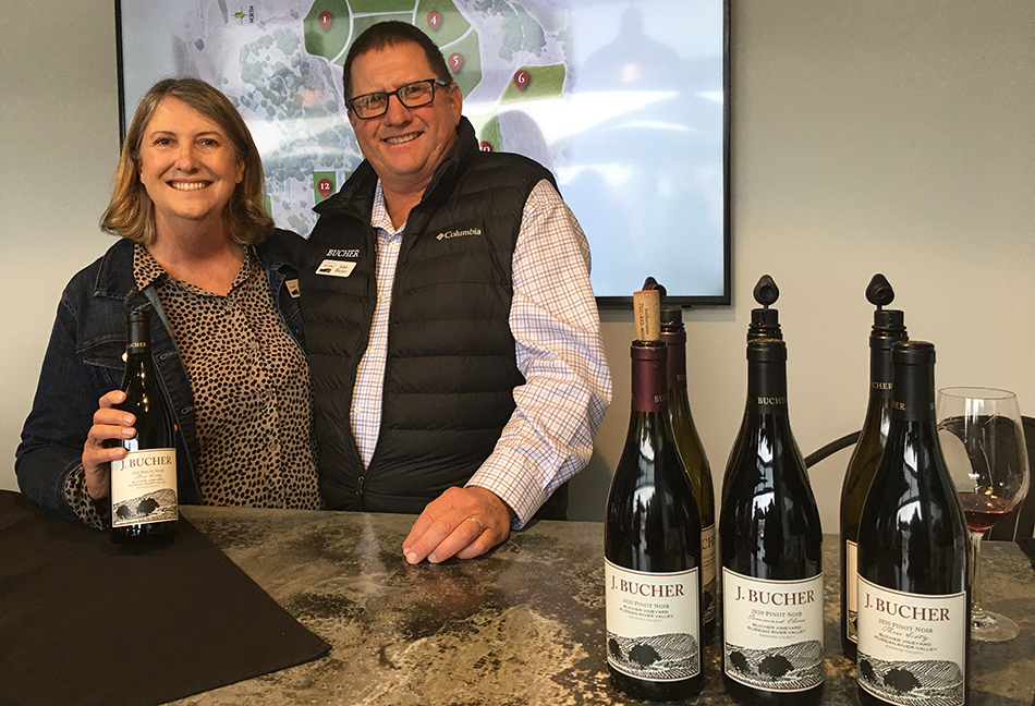 Diane and John Bucher with Bucher Wine bottles on a counter and a map of their vineyard in the background.