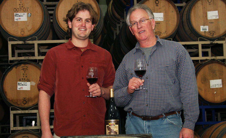 Jamie and Fred Peterson holding wine glasses, standing in front of stacks of barrels.