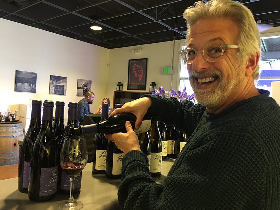 Jeff Cohen pouring wine into a Wine Road logo glass with the wine bar covered with Jeff Cohen wine bottles.