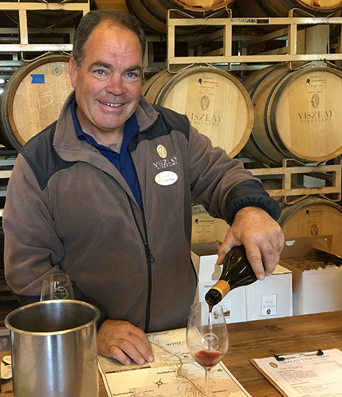 John Viszlay pouring wine into a Wine Road logo glass with barrels in the background.