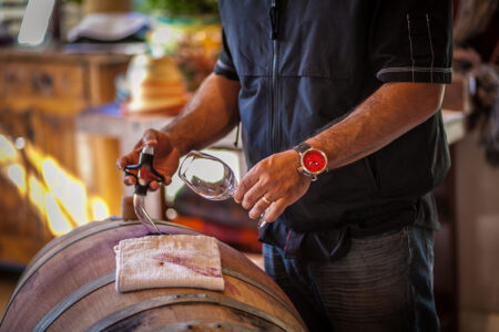Man, all dressed in blue with a red-faced watch, has a wine thief inserted in a wine barrel in one hand and a wine glass in the other hand.