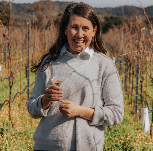 Winemaker Ashely Herzberg standing in a vineyard in winter. Holding a glass of wine and the vineyard rows have orange flowering plants as the cover crop.