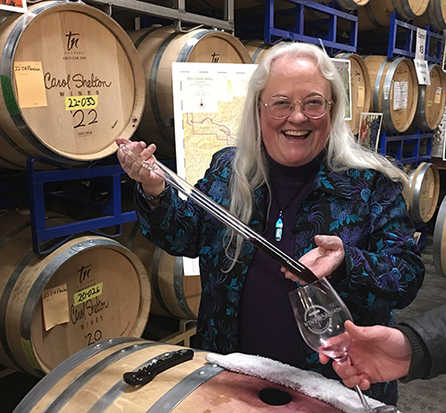 Carol Shelton pouring wine from a barrel using a wine thief into a Wine Road wine glass. Barrels in the background with Carol Shelton Wines stamped on each barrel.