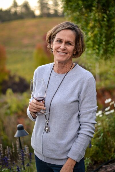 Diane Wilson standing outside, holding a glass with red wine, with muted green colors in the background.