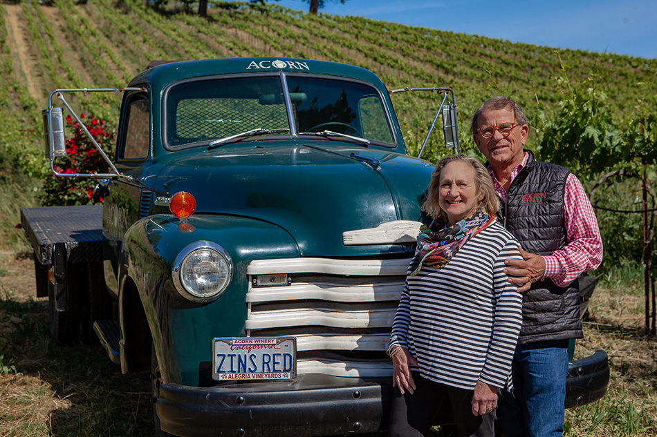 A man and woman stand in front of an old green-cabbed truck with hillside vineyards in the background. The truck's licenses plate reads ZINS RED.