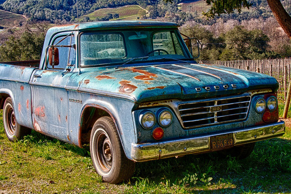 Old rusty blue Dodge pickup truck sitting next to a vineyard in spring. Hillside of trees and vineyards in the background.