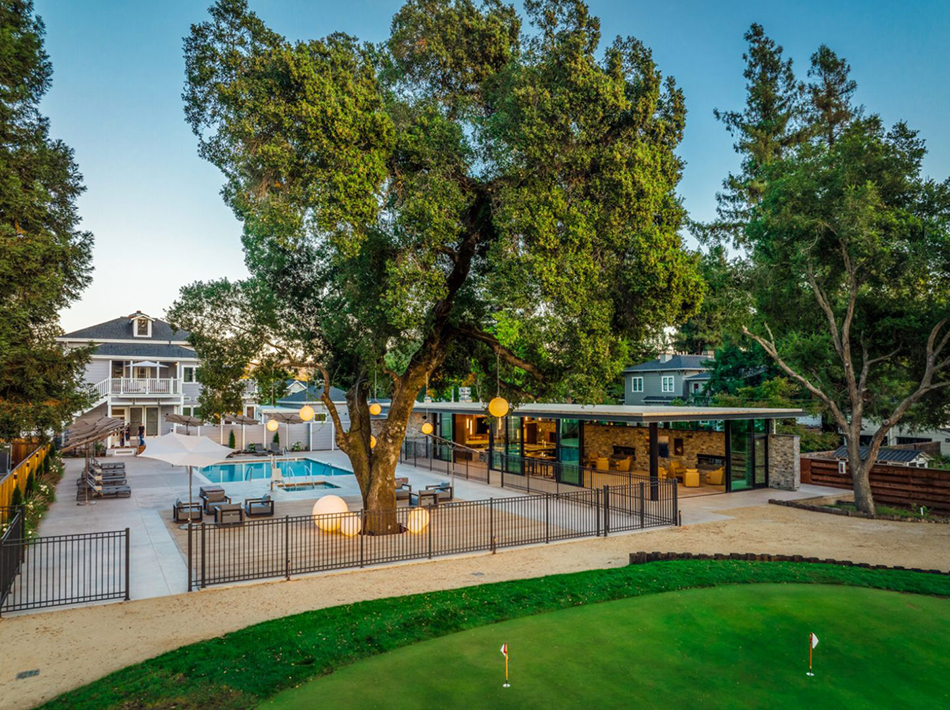 A view of an expansive backyard with mature trees. At the far end is a large house. In the middle is a large deck surrounding a pool. Off to the right side is a partially enclosed seating and eating area. At the close end is a putting green.