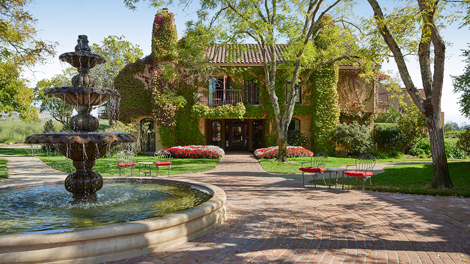 Three tiered fountain with water spilling over. Brick laid walking paths with chairs and tables off to the sides. A Mediterranean style two-story building at the end of the path. Mature trees, flower beds and green grass fill in the landscape.