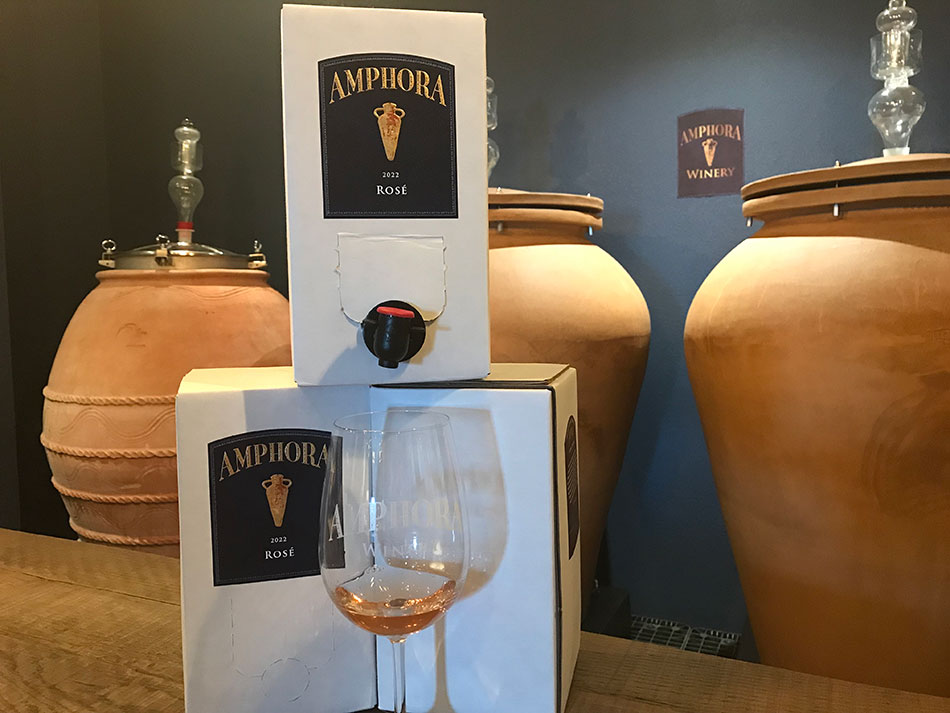 In the foreground is a wine glass with the Amphora logo, and has a small amount of rose colored wine in it. Three boxes of wine with Amphora labels are right behind the wine glass. In the background are clay amphora pots used to ferment wine.