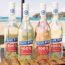 Four Blue Bin wine bottles with labels that read 100% recycled bottle and the name of the wine. The background is a beach and ocean with boats in the bay and buildings on the hills on the right side.