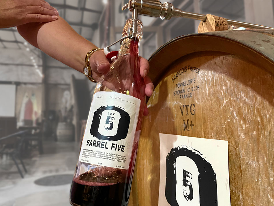 A hand is holding a growler bottles with a Barrel 5 label on it. The growler is being filled from a French oak wine barrel through a spigot.