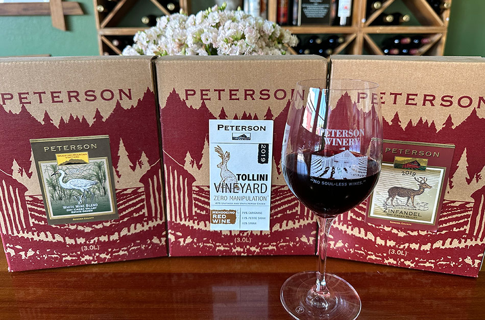A glass of red wine with a Peterson Winery logo. Behind are three boxes of Peterson wine with colorful labels in the center of each box.