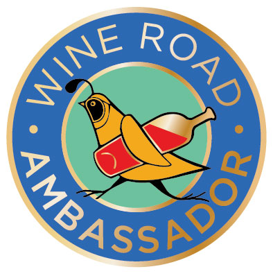 a circle with a stylized quail holding a wine bottle. Around the edges of the circle are the words Wine Road Ambassador